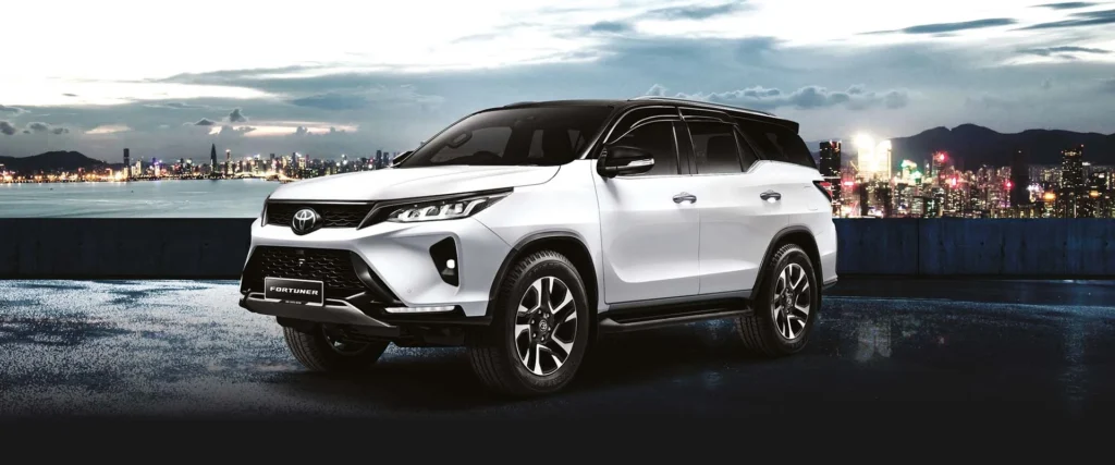 White Toyota Fortuner SUV positioned against urban waterfront backdrop with dramatic cityscape and mountain silhouette