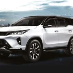 White Toyota Fortuner SUV positioned against urban waterfront backdrop with dramatic cityscape and mountain silhouette