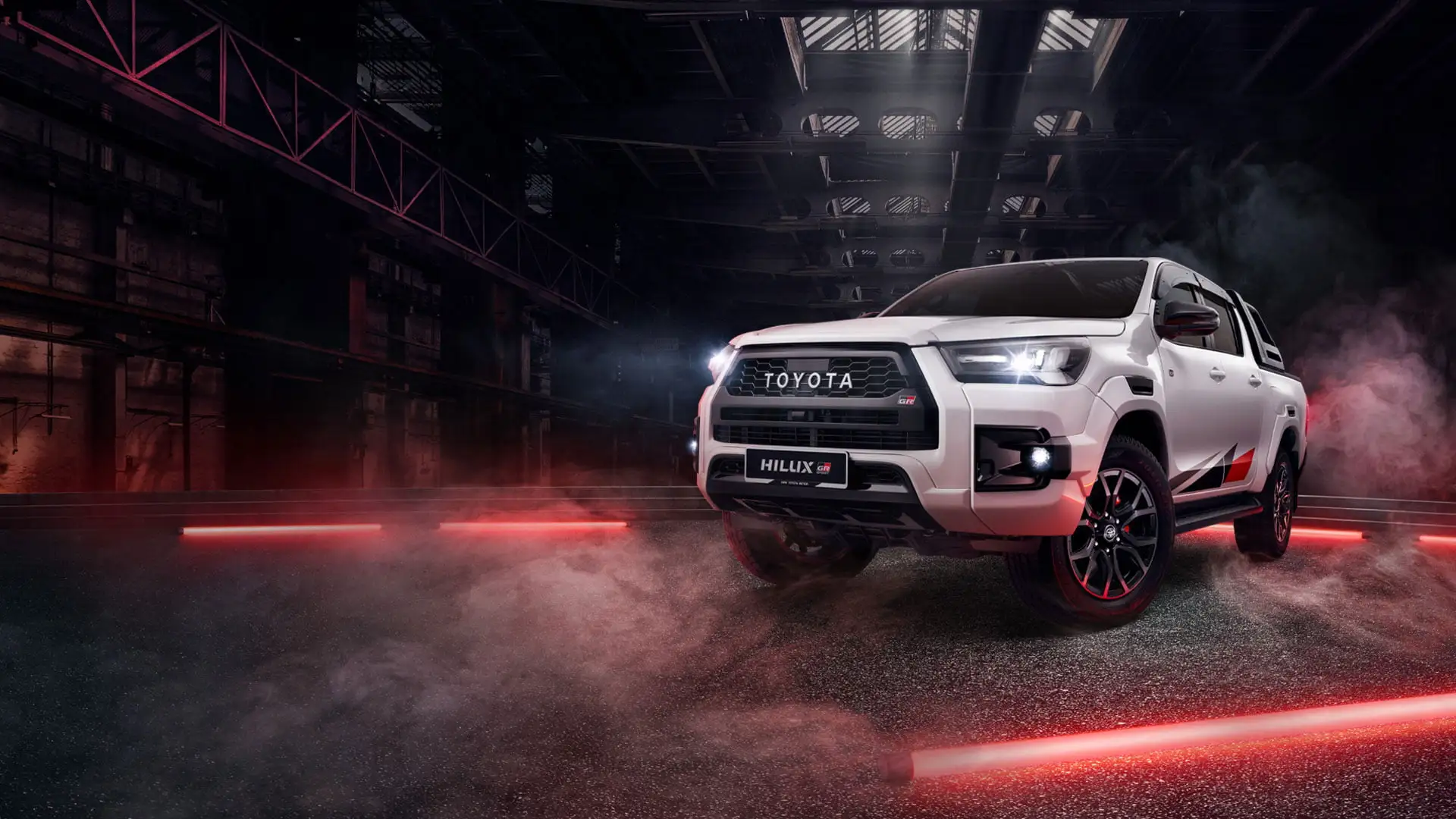 Toyota Hilux GR Sport featuring aggressive front grille and black wheels in atmospheric industrial setting with red accents