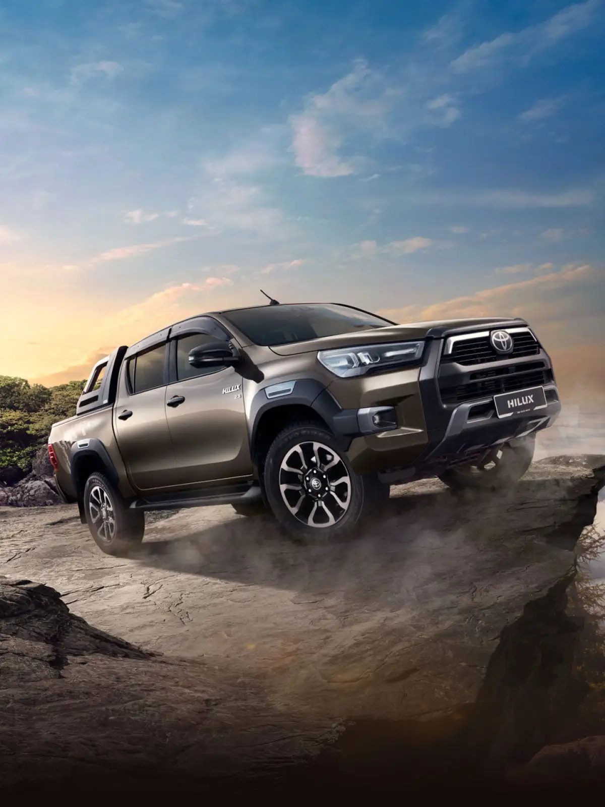 Bronze Toyota Hilux pickup truck positioned on rocky mountain terrain with dramatic sunset and rugged landscape backdrop