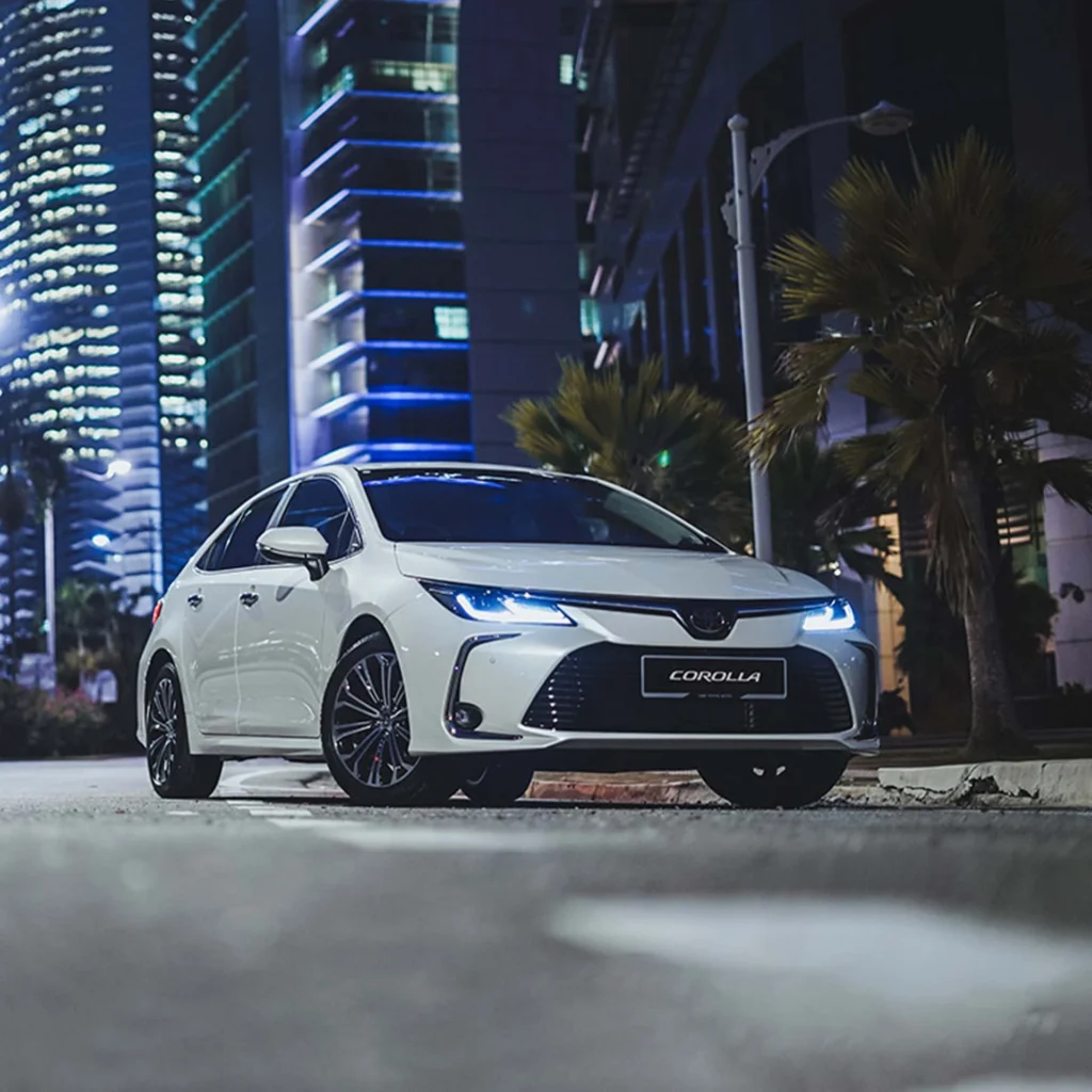 White Toyota Corolla at night with LED headlights on urban street with illuminated skyscrapers and palm trees