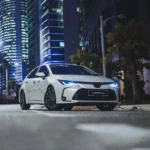 White Toyota Corolla at night with LED headlights on urban street with illuminated skyscrapers and palm trees