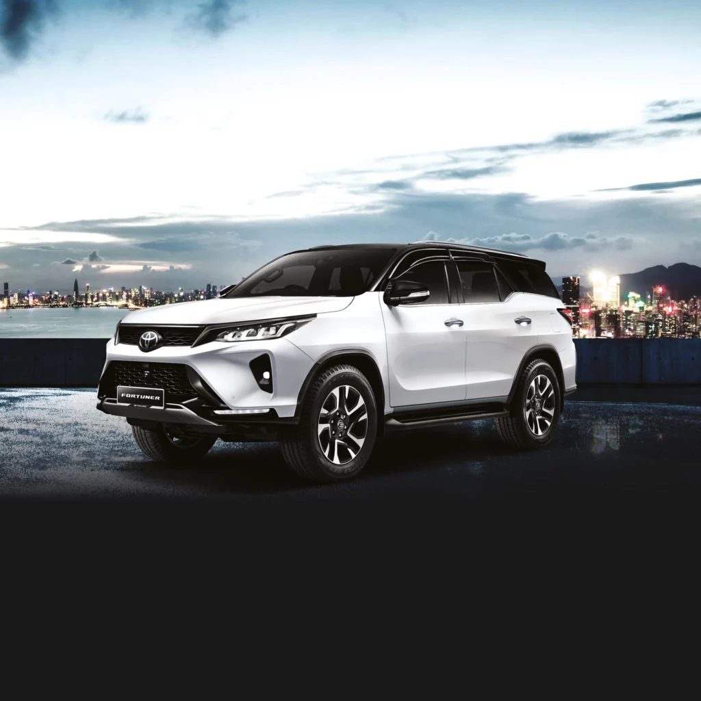 White Toyota Fortuner SUV positioned against urban waterfront backdrop with dramatic cityscape and mountain silhouette