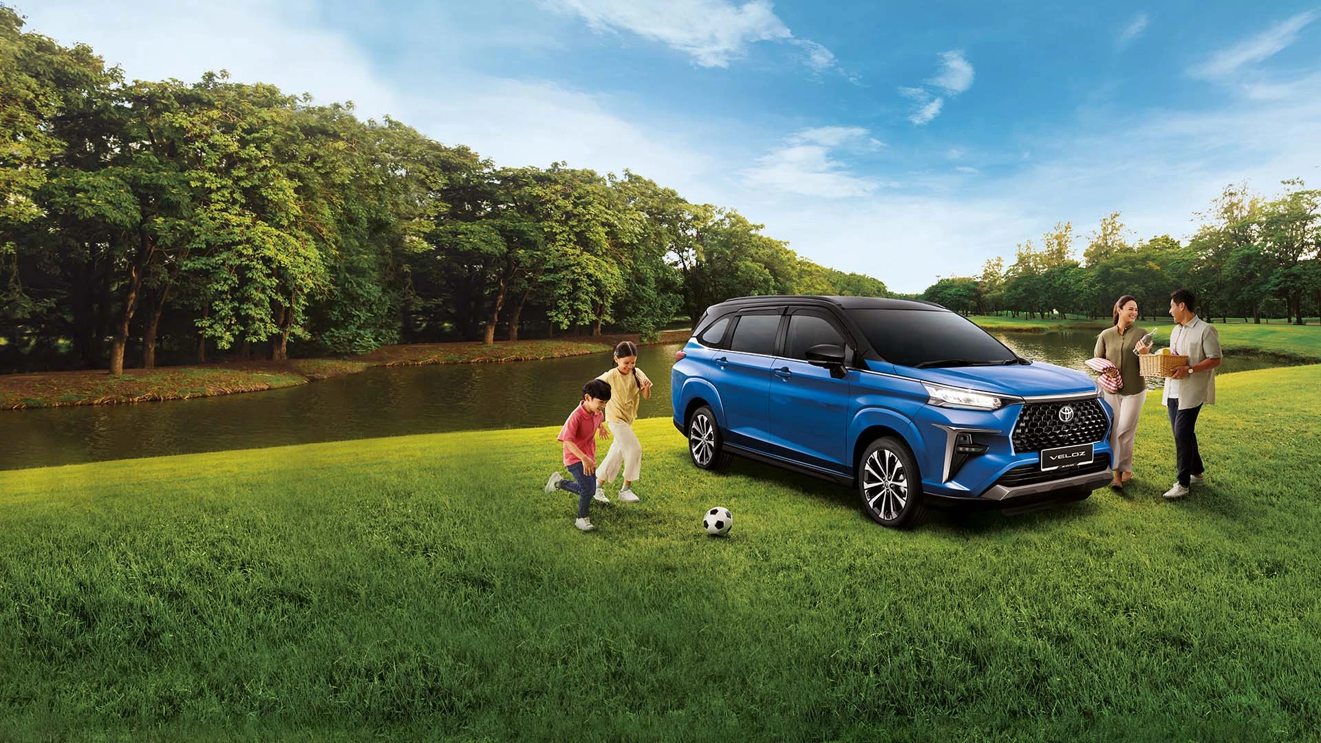 Toyota Veloz blue parked on green grass beside lake with family enjoying outdoor picnic and children playing