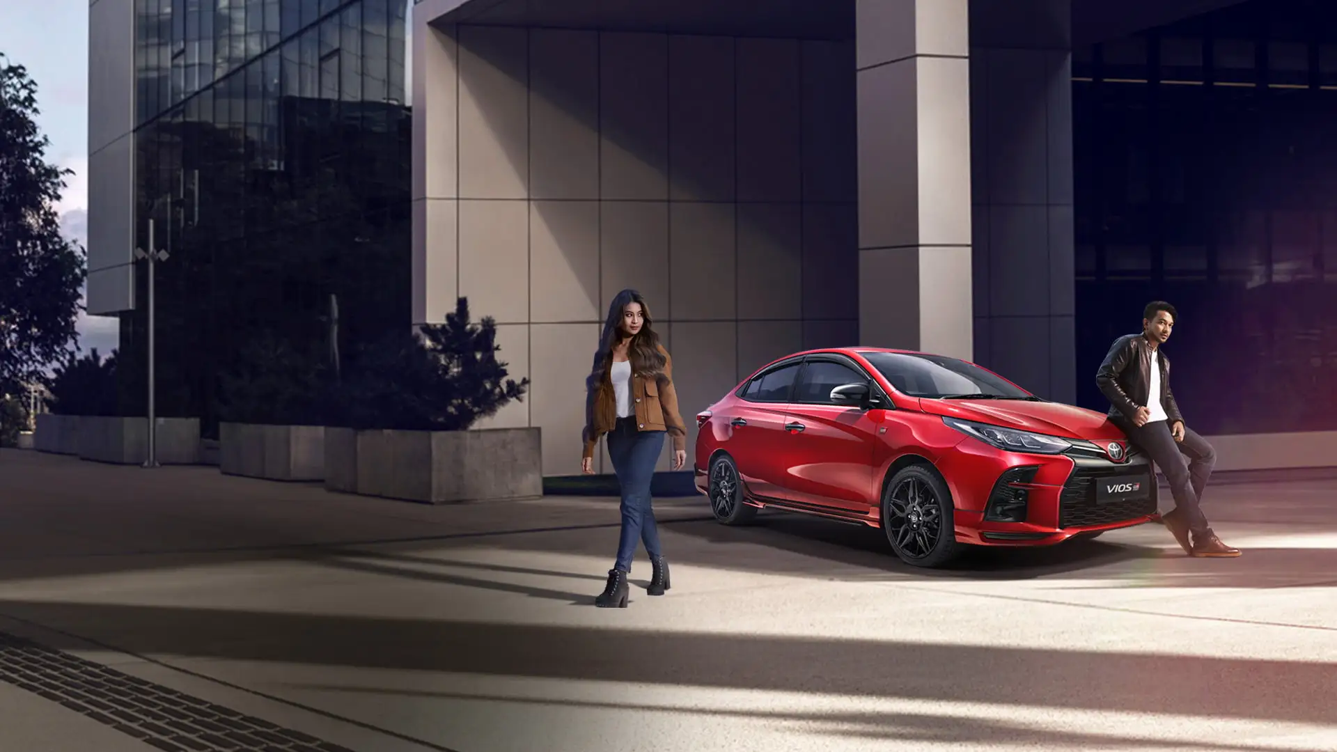 Red Toyota Vios sedan with stylish couple posing near modern glass building during golden hour lighting