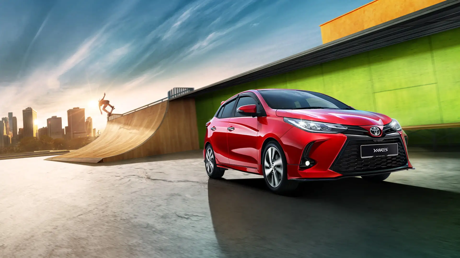 Toyota Yaris in red positioned near skateboarding ramp with modern cityscape and dramatic sky during golden hour