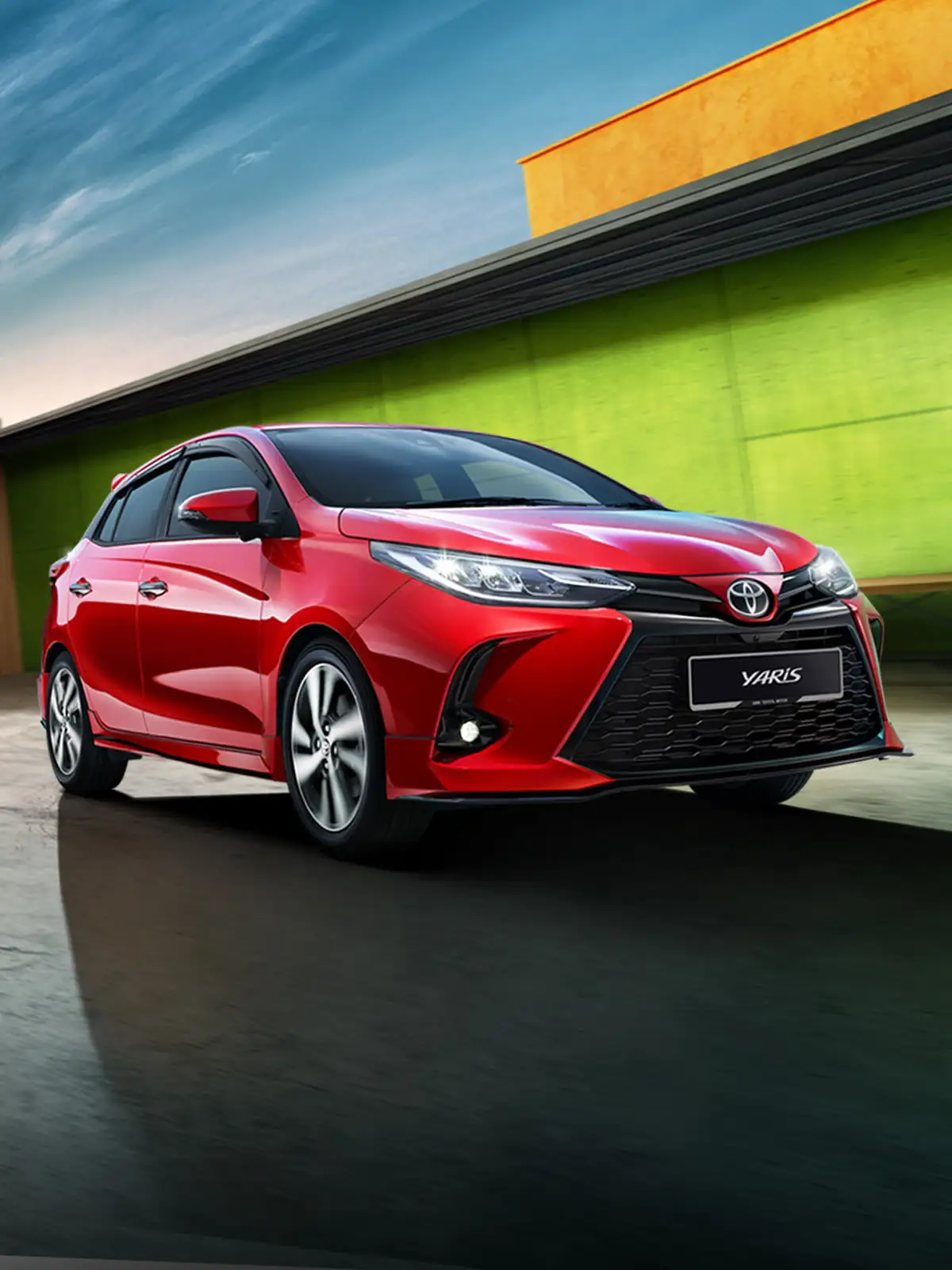 Toyota Yaris in red positioned near skateboarding ramp with modern cityscape and dramatic sky during golden hour
