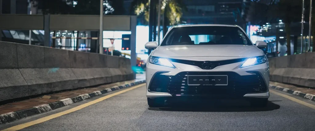 Toyota Camry facing forward in nighttime urban setting with illuminated LED headlights
