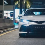 Toyota Camry facing forward in nighttime urban setting with illuminated LED headlights